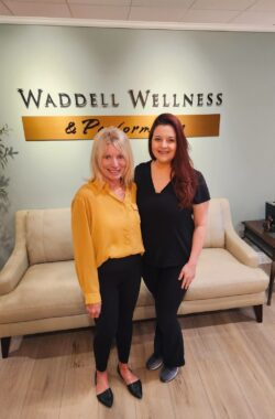 Two women stand side by side and smile for a photo. The wall behind them has a sign that reads "Waddell Wellness and Performance."