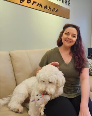 A young woman sits on a couch in a chiropractic office and pets a fluffy white dog.