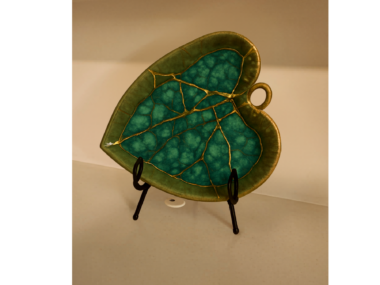 A two-toned green marble pottery plate in the shape of a heart sits on a stand. It is covered in golden cracks.