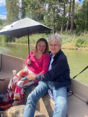 A couple sits in a small boat that is floating down a river in the Netherlands. The woman is laughing and holding up an umbrella to block the sun, while the man is smiling and has his arm around his wife.